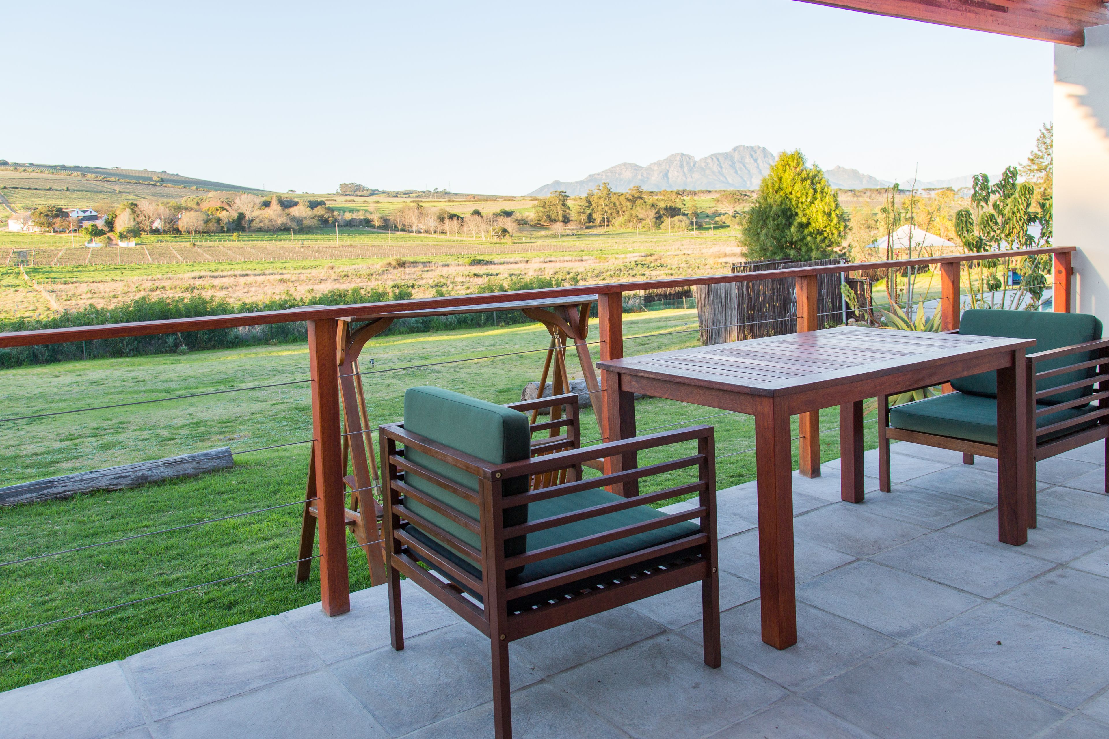 A patio with a wooden table and two chairs, overlooking a green landscape with mountains in the distance.