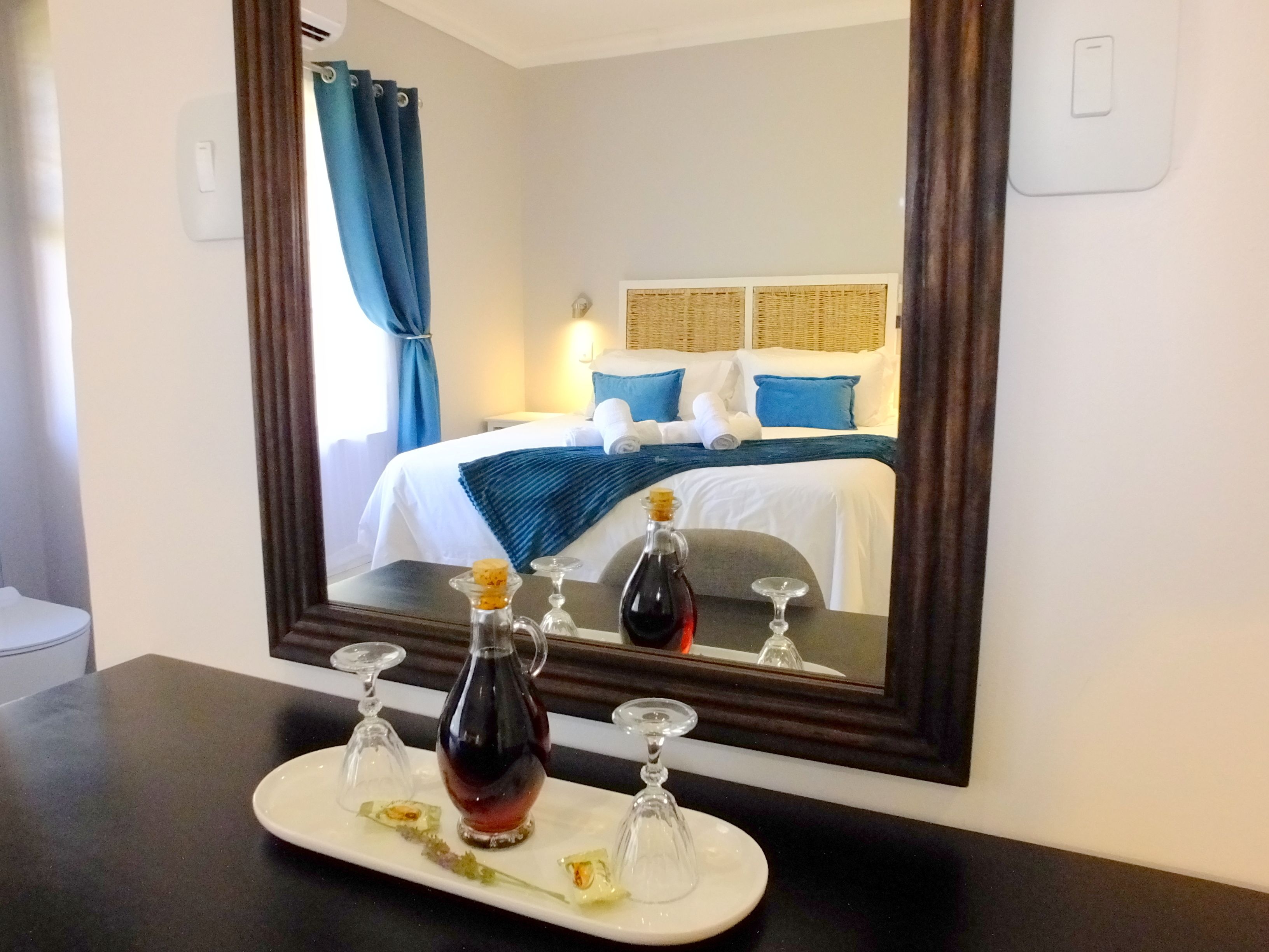 A hotel room with a bed, a bedside table with a bottle and glasses, and a mirror reflecting the room.