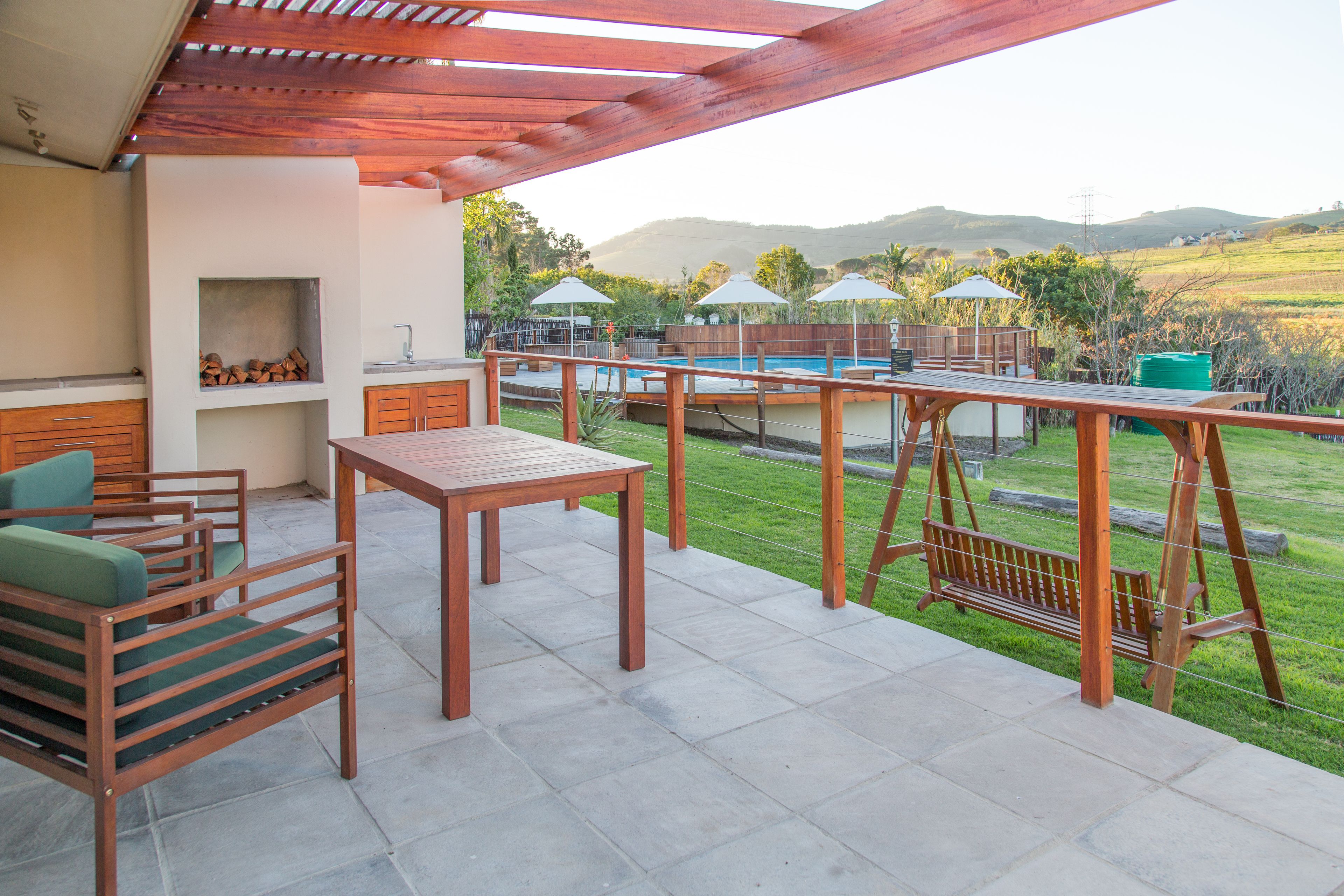 Une terrasse extérieure avec une table et des chaises en bois, un espace barbecue et une vue sur une piscine et des collines en arrière-plan.