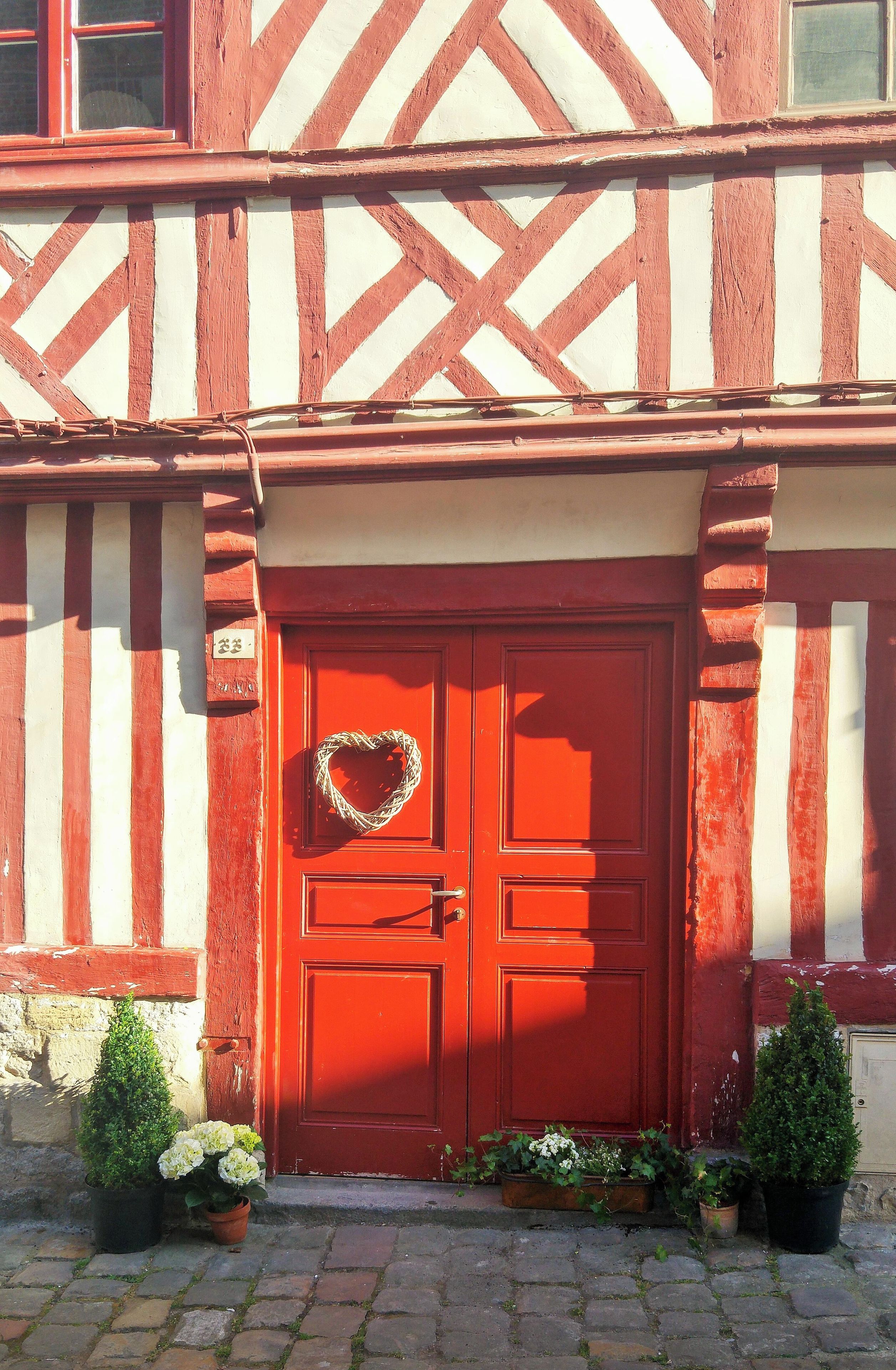 Une porte rouge ornée d’un motif en forme de cœur, flanquée de plantes en pot et donnant sur une entrée pavée de pierres.