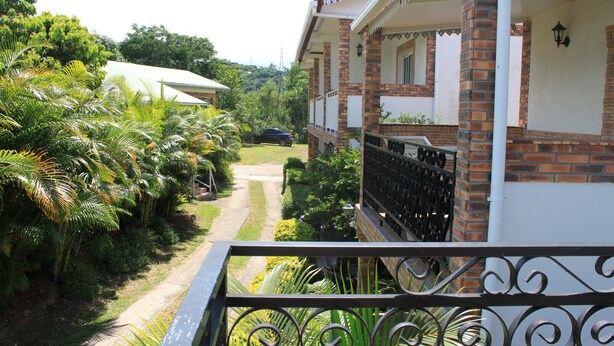 A residential building with a brick facade, a balcony with decorative metal railings, and lush greenery surrounding the property.