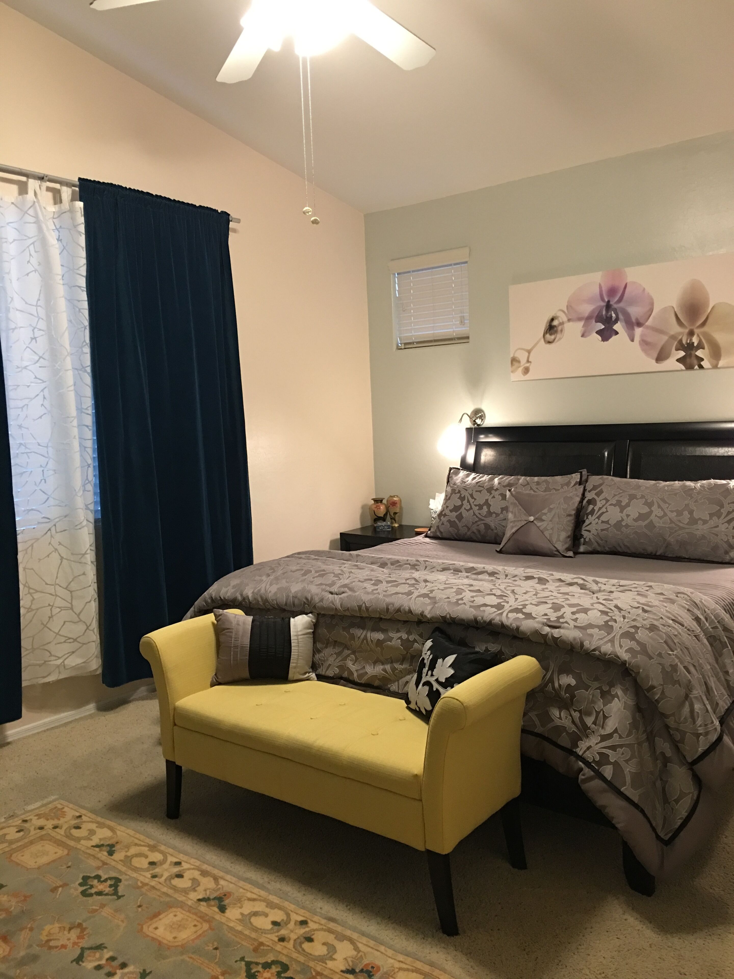 A bedroom with a bed, a nightstand, a lamp, a chair, a window with blinds, and a ceiling fan.
