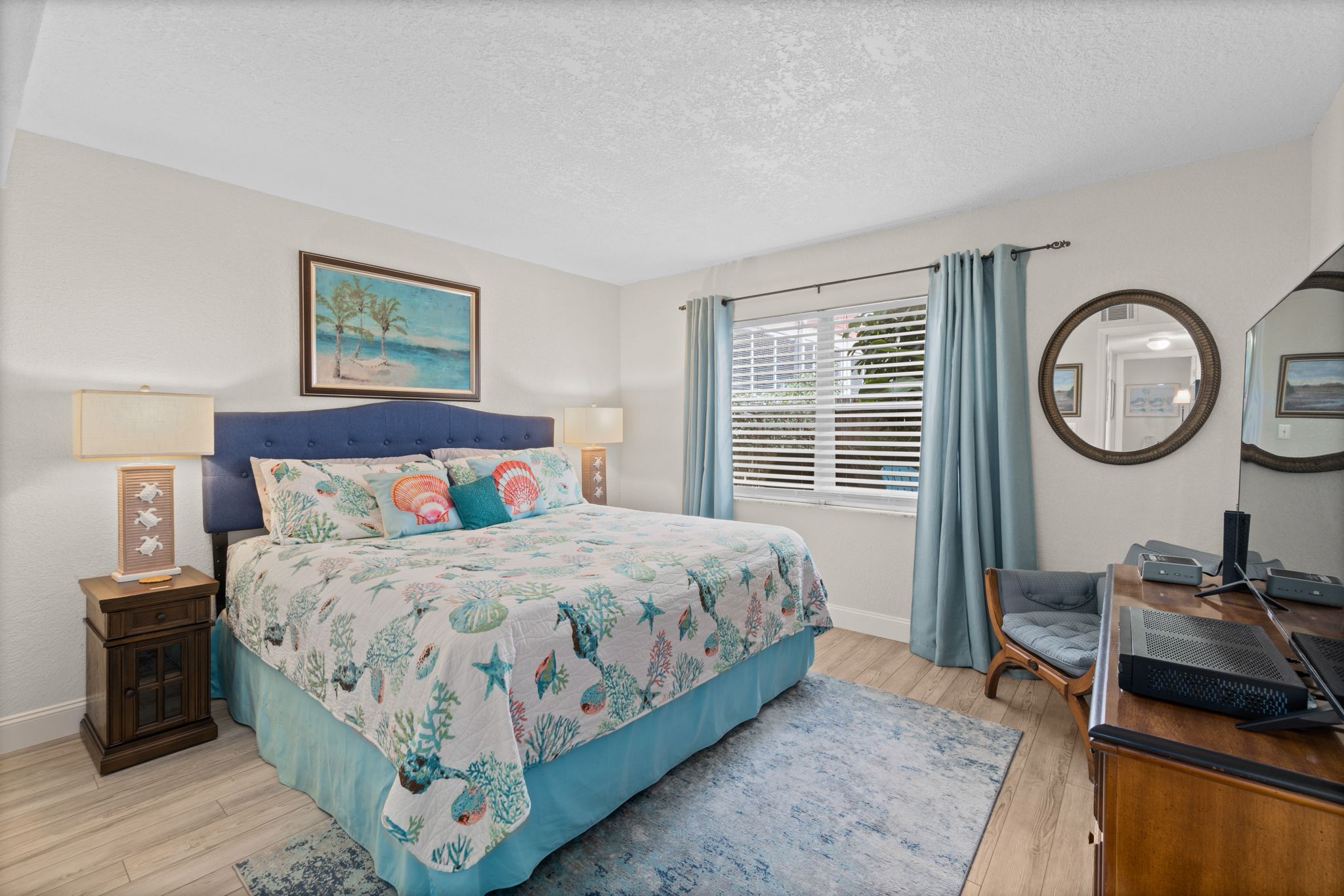 A bedroom with a bed, a desk, a chair, a window with curtains, a nightstand, a lamp, and a painting on the wall.
