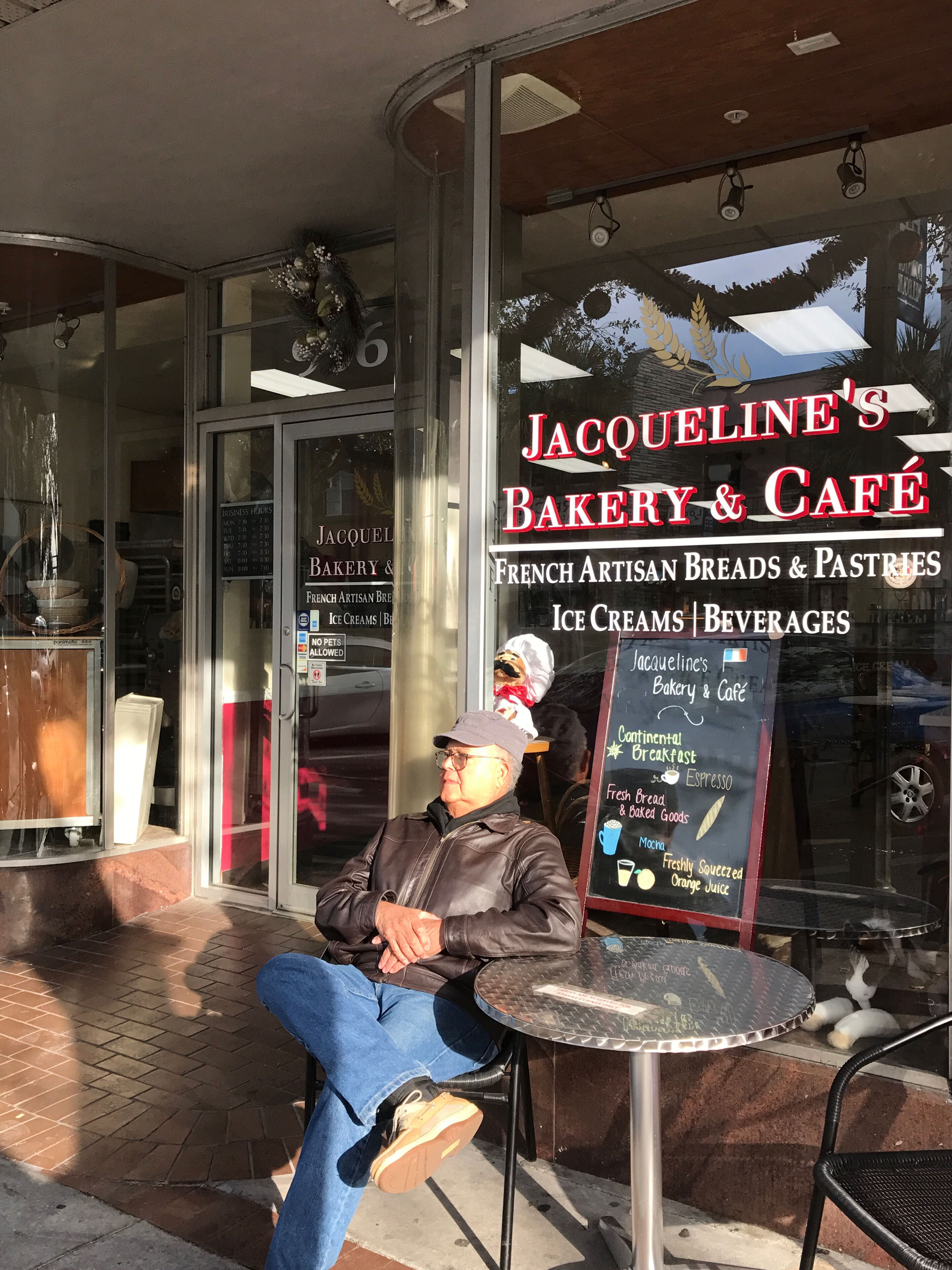 Un área de estar al aire libre frente a Jacqueline's Bakery & Café, con un panel de menú a la vista.