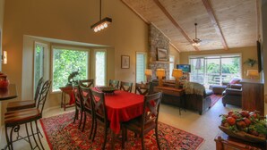 A dining area with a red tablecloth, wooden chairs, and a stone wall. There is a stone fireplace, a ceiling fan, and a large window with blinds. - Townhome w/ Mtn Views & Club Access! Indoor Pool, Near Blowing Rock & Skiing (Boone)
