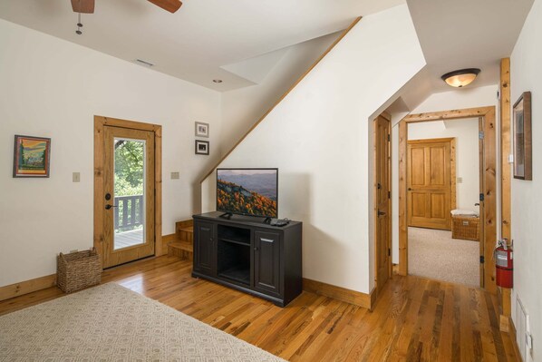 A spacious room with wooden flooring, a television on a cabinet, a staircase, and a door leading outside.