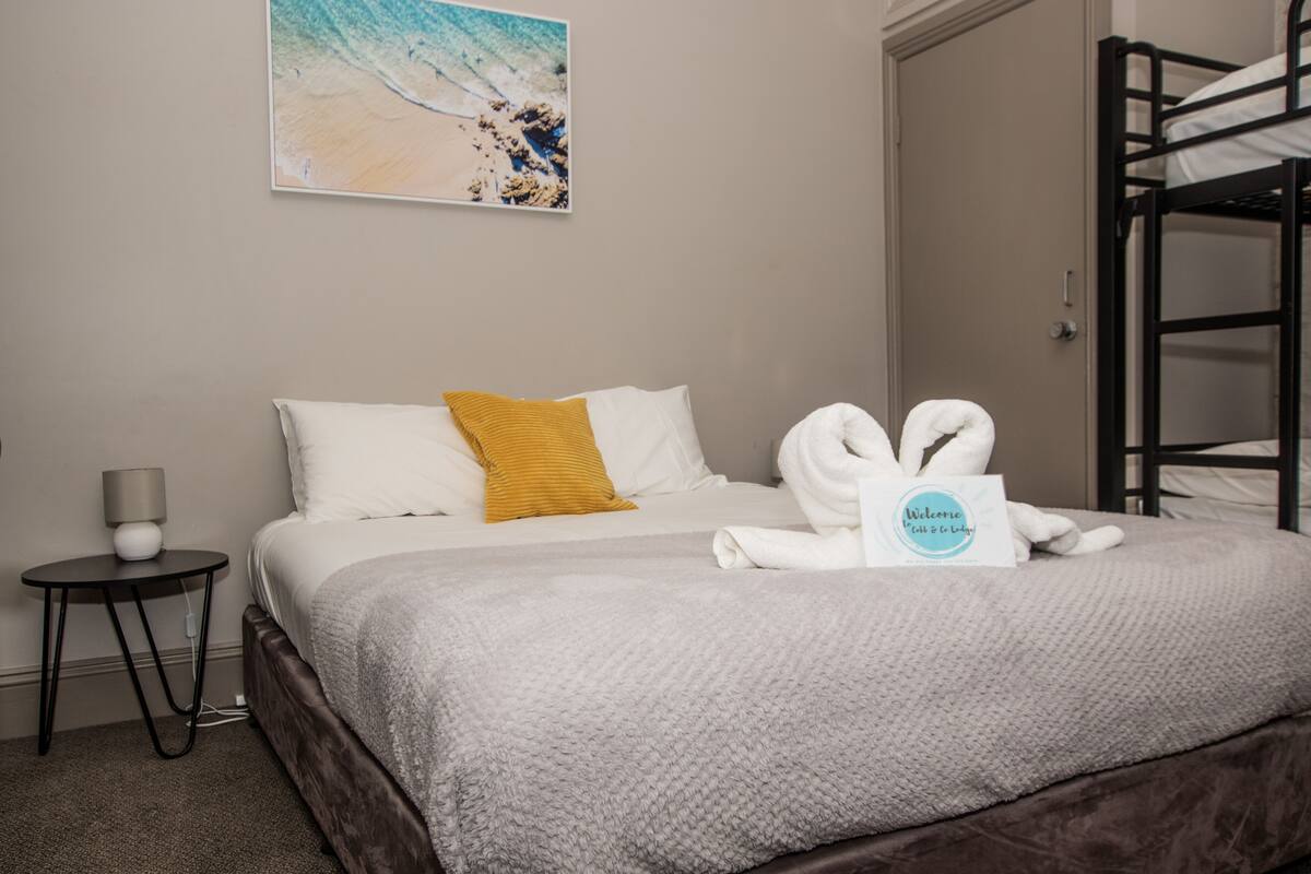 A neatly made bed with a gray blanket, a white pillow, and a mustard-colored pillow. There is a welcome mat with a logo on the bed.