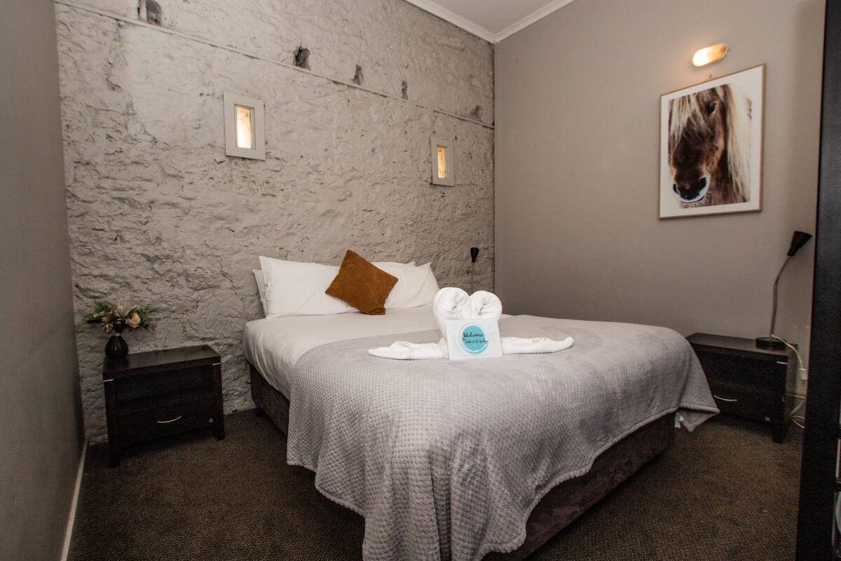 A hotel room with a stone-textured wall, a bed with a gray blanket, a brown pillow, and a framed picture of a horse on the wall.
