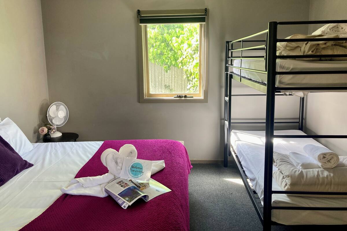 A hotel room with a bed, a window with a view of greenery, a bunk bed, a bedside table with a fan, and a welcome card.