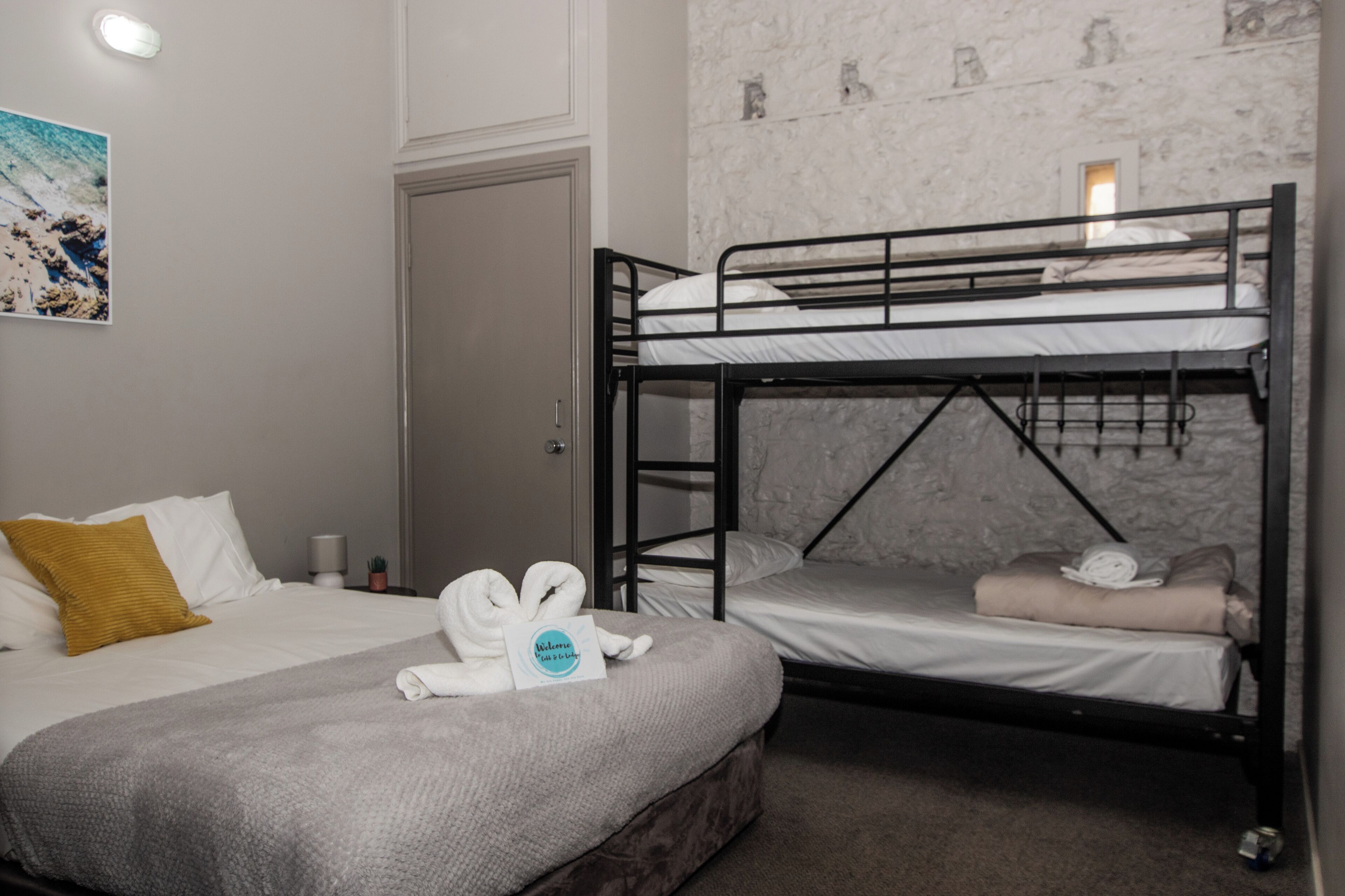 Hotellihuone, jossa on kerrossänky, yksi erillinen sänky, harmaa peitto, tervetulotoivotus ja taulu seinällä.