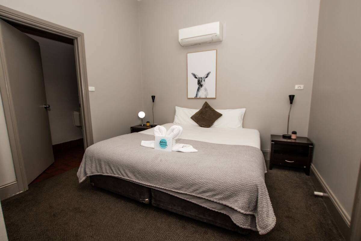 A hotel room with a bed, a nightstand, a lamp, an air conditioner, and a framed picture on the wall.
