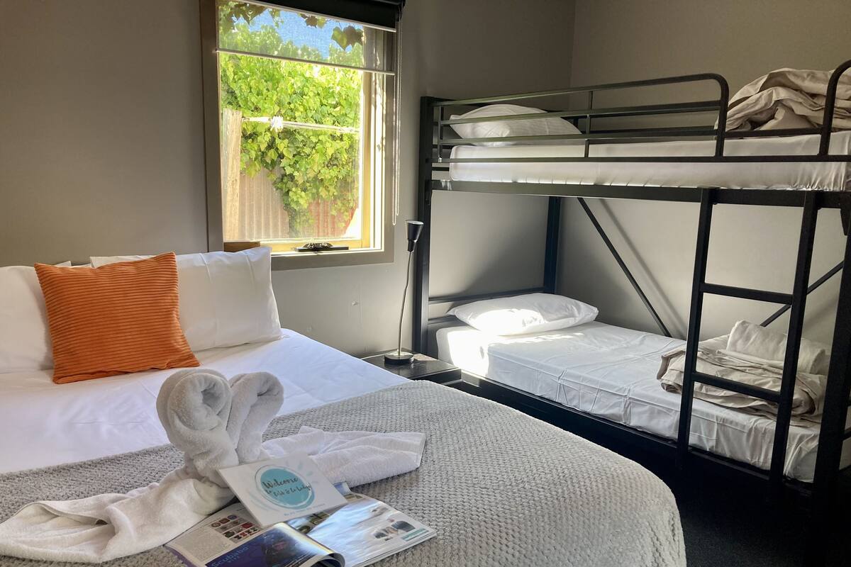 A hotel room with a bunk bed, a single bed, a window with a view of greenery, a towel, and a welcome booklet.