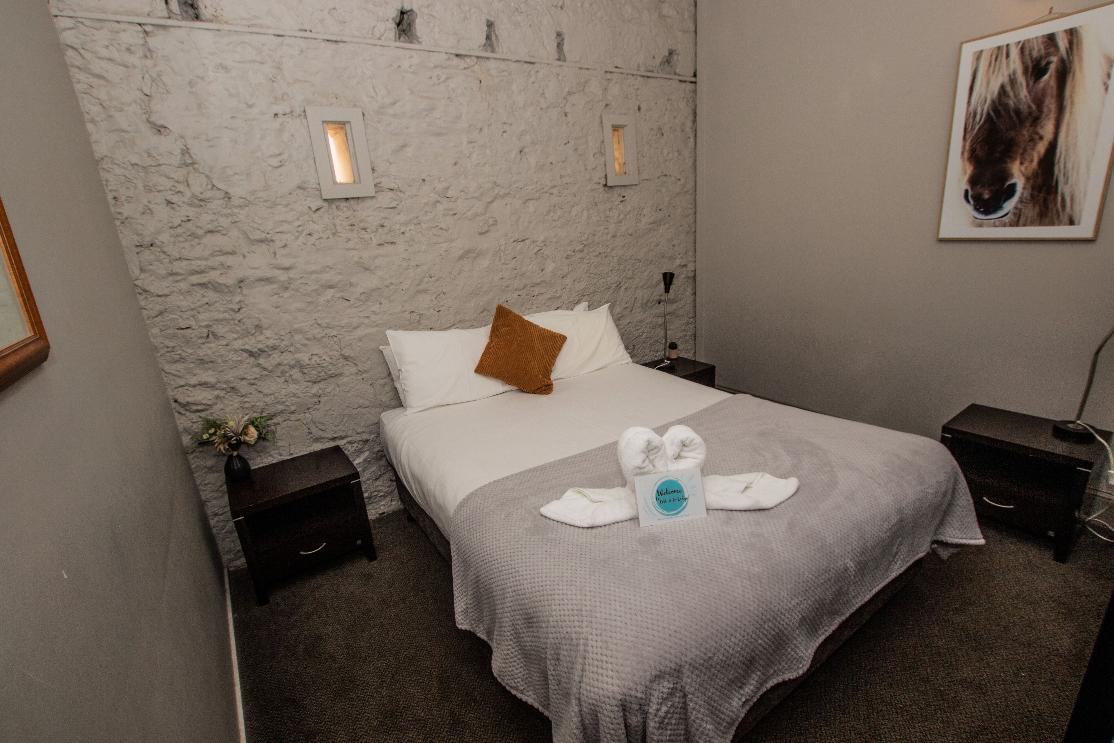 A hotel room with a marble wall, a bed with white and brown pillows, a towel and a sign on the bed, a framed picture on the wall, and a small plant on the nightstand.