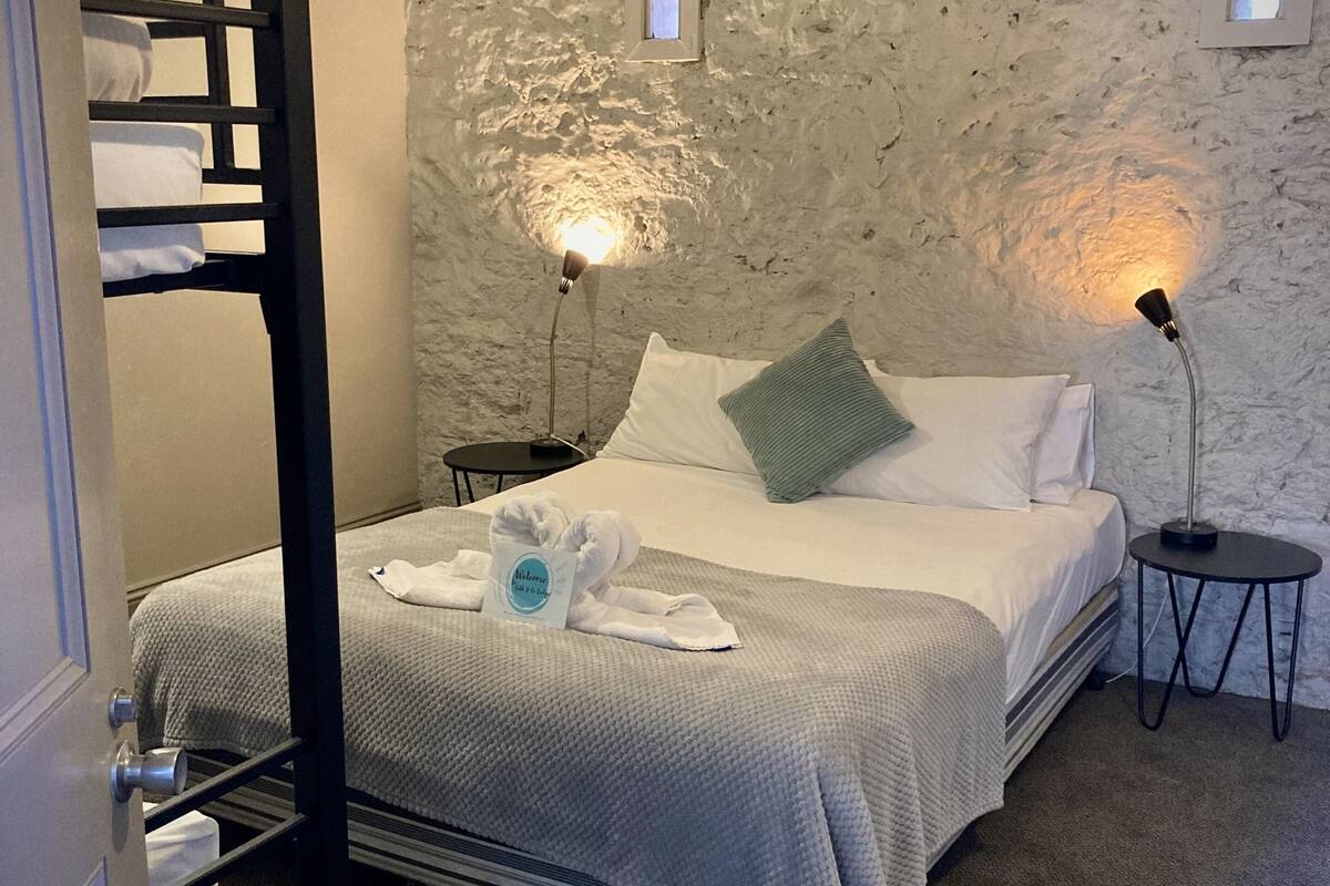A single bed with a grey quilt, a green pillow, and a towel on the floor. There are two bedside tables with lamps. The walls are textured and there are small windows.