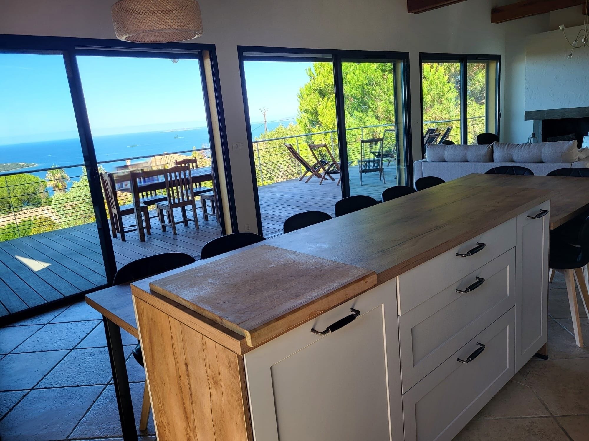 A modern kitchen with a wooden countertop and white cabinets, overlooking a deck with outdoor furniture and a view of the ocean.