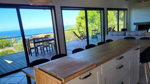 A modern kitchen with a wooden countertop and white cabinets, overlooking a deck with outdoor furniture and a view of the ocean.