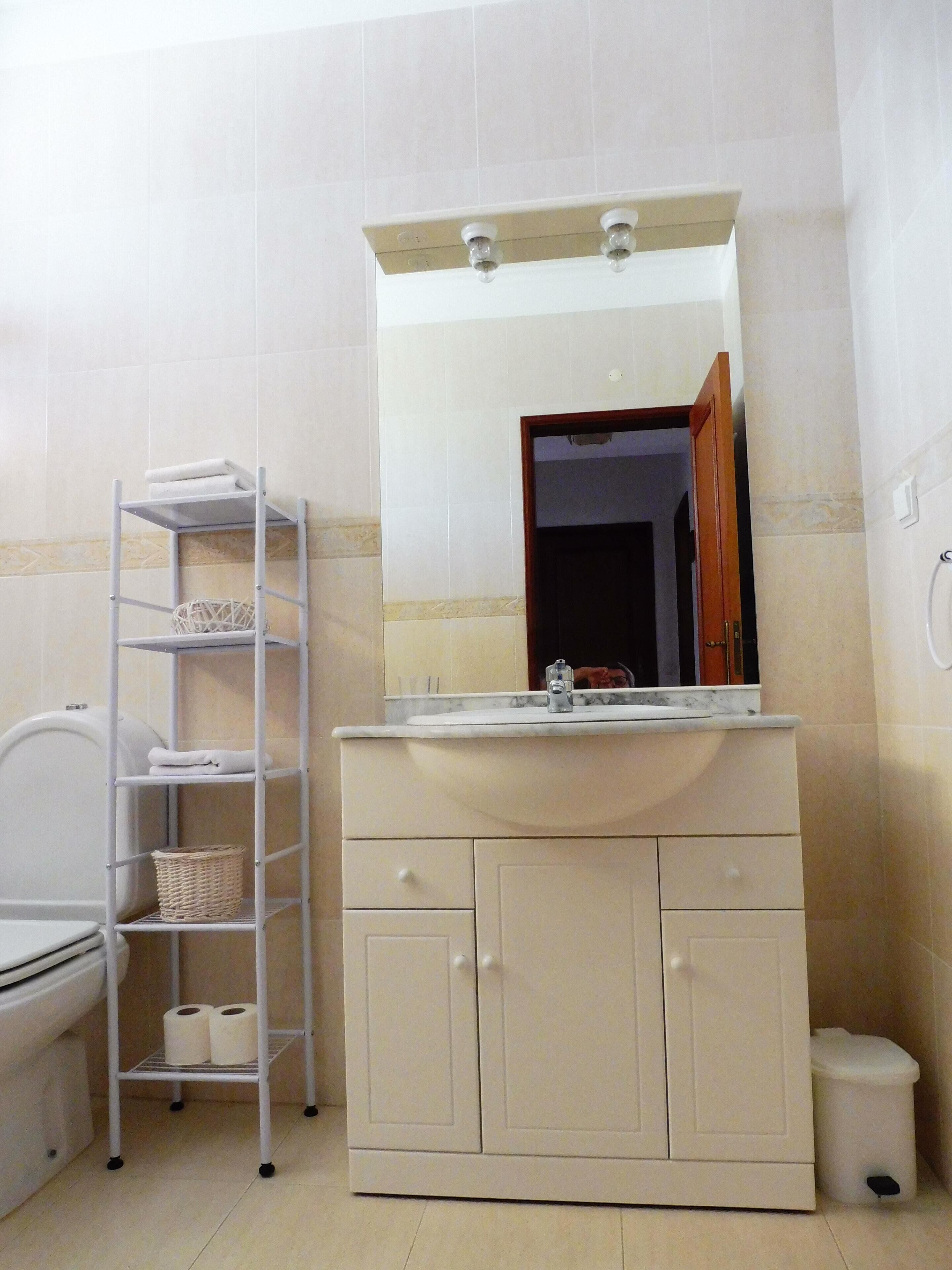 Une salle de bain avec un meuble-lavabo blanc, un miroir au-dessus du lavabo, des toilettes et une étagère métallique avec des paniers et des serviettes.