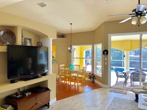 A living room with a flat-screen TV, wooden entertainment center, dining area with yellow chairs, and a balcony with outdoor furniture. - Southern Exposure, Gulf Access, Heated Pool Home with a Hot Tub and Large Lanai! (Cape Coral)