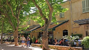 An outdoor café area with tables and chairs under tree canopies, surrounded by buildings with signage indicating the names of the establishments. - Saint Paul De Vence, Charming Suites in The Heart Of The Village. 4 ** Biloba (Saint-Paul-De-Vence)