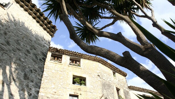 A stone building with a tiled roof, multiple windows, and a large tree in front. - Saint Paul De Vence, Charming Suites in The Heart Of The Village. 4 ** Biloba (Saint-Paul-De-Vence)