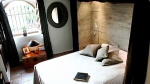 A bedroom with a bed, a bedside table, a mirror, and a window with a view of greenery. - Saint Paul De Vence, Charming Suites in The Heart Of The Village. 4 ** Biloba (Saint-Paul-De-Vence)