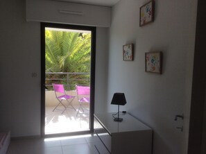 Une pièce dotée d’une porte-fenêtre coulissante donnant sur un balcon agrémenté d’un palmier et de deux chaises roses. Trois tableaux encadrés ornent le mur.