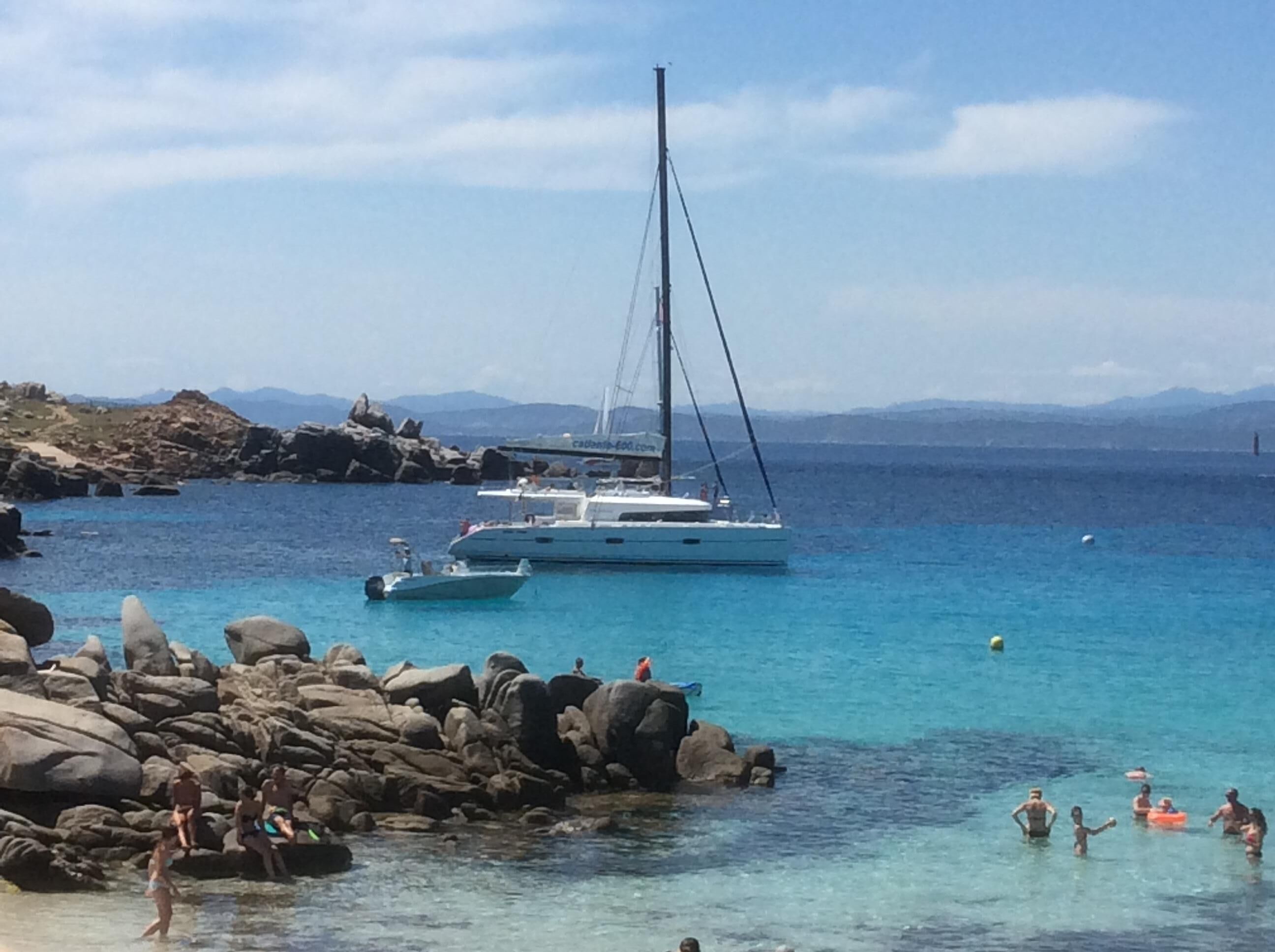 A rocky beach with clear turquoise water, a sailboat anchored nearby, and people enjoying the water.