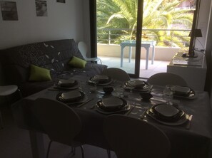 Uma sala de jantar com mesa posta para quatro pessoas, um sofá com almofadas e vista para palmeiras através de grandes janelas.