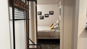 Kompakti hotellihuone, jossa on sänky, pieni oleskelualue penkillä sekä seinälle kiinnitetty hylly, jossa on kasvi.
