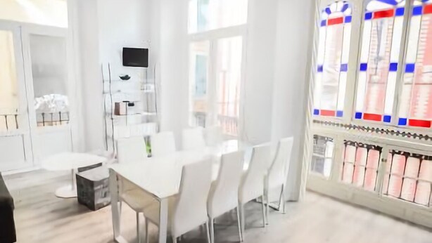 Une pièce moderne aux tons clairs, avec un espace repas comprenant une longue table et des chaises, un téléviseur fixé au mur et une grande fenêtre ornée de vitraux.