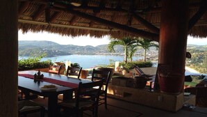 An outdoor dining area with a table and chairs, a thatched roof, and a view of a lake and mountains.