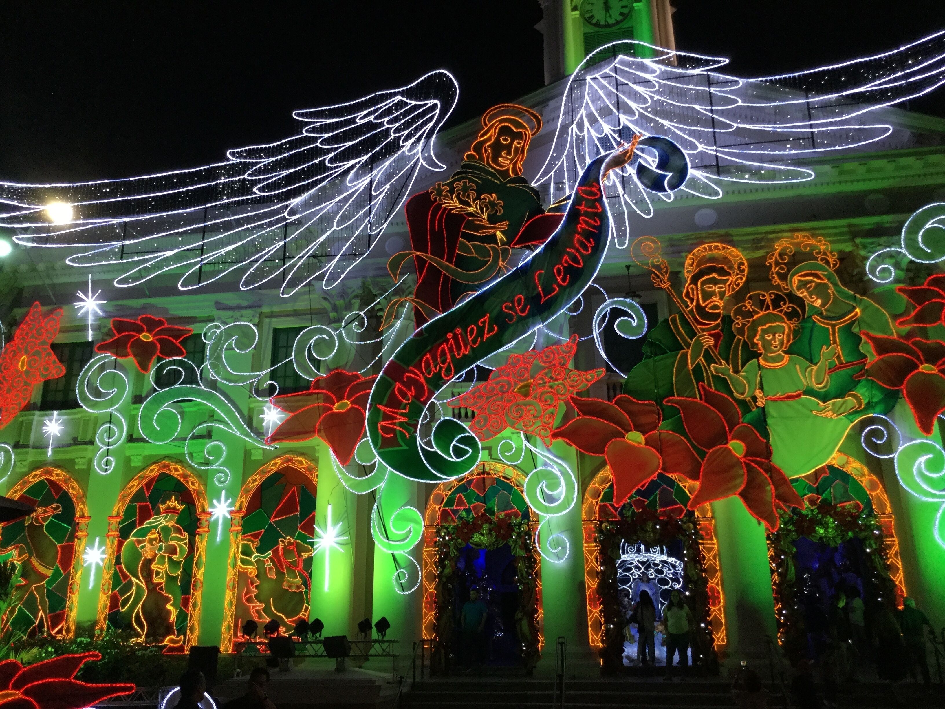 A festive outdoor display with illuminated figures and decorations, including a prominent winged figure and a banner reading "Magiiez se Levi."