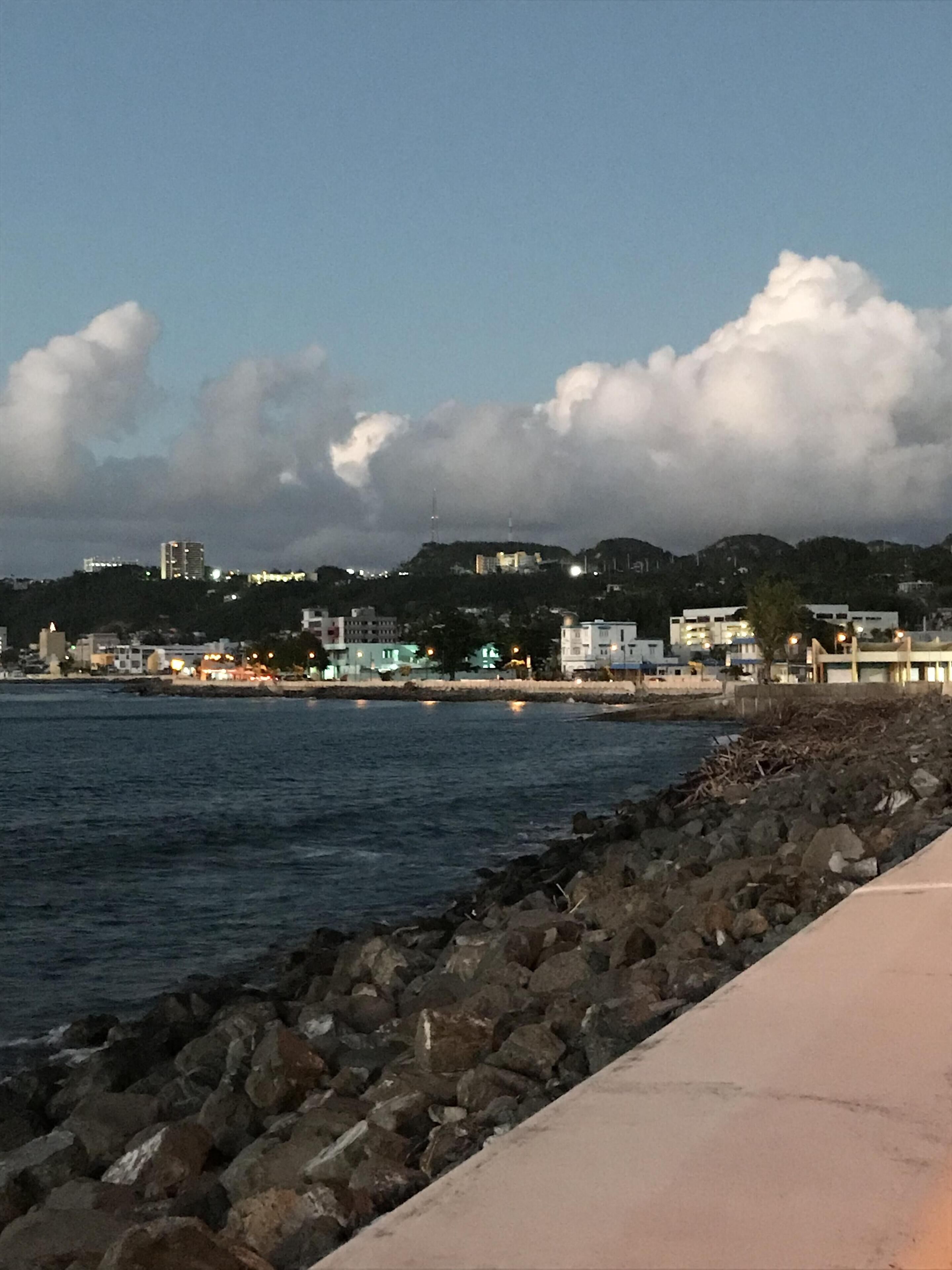 Vista costera con costa rocosa, un paseo pavimentado y edificios al fondo bajo un cielo parcialmente nublado.