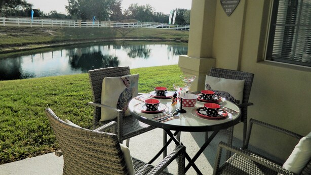 Un patio con sillas de mimbre y una mesa para dos, con vista a un lago tranquilo y una cerca blanca.