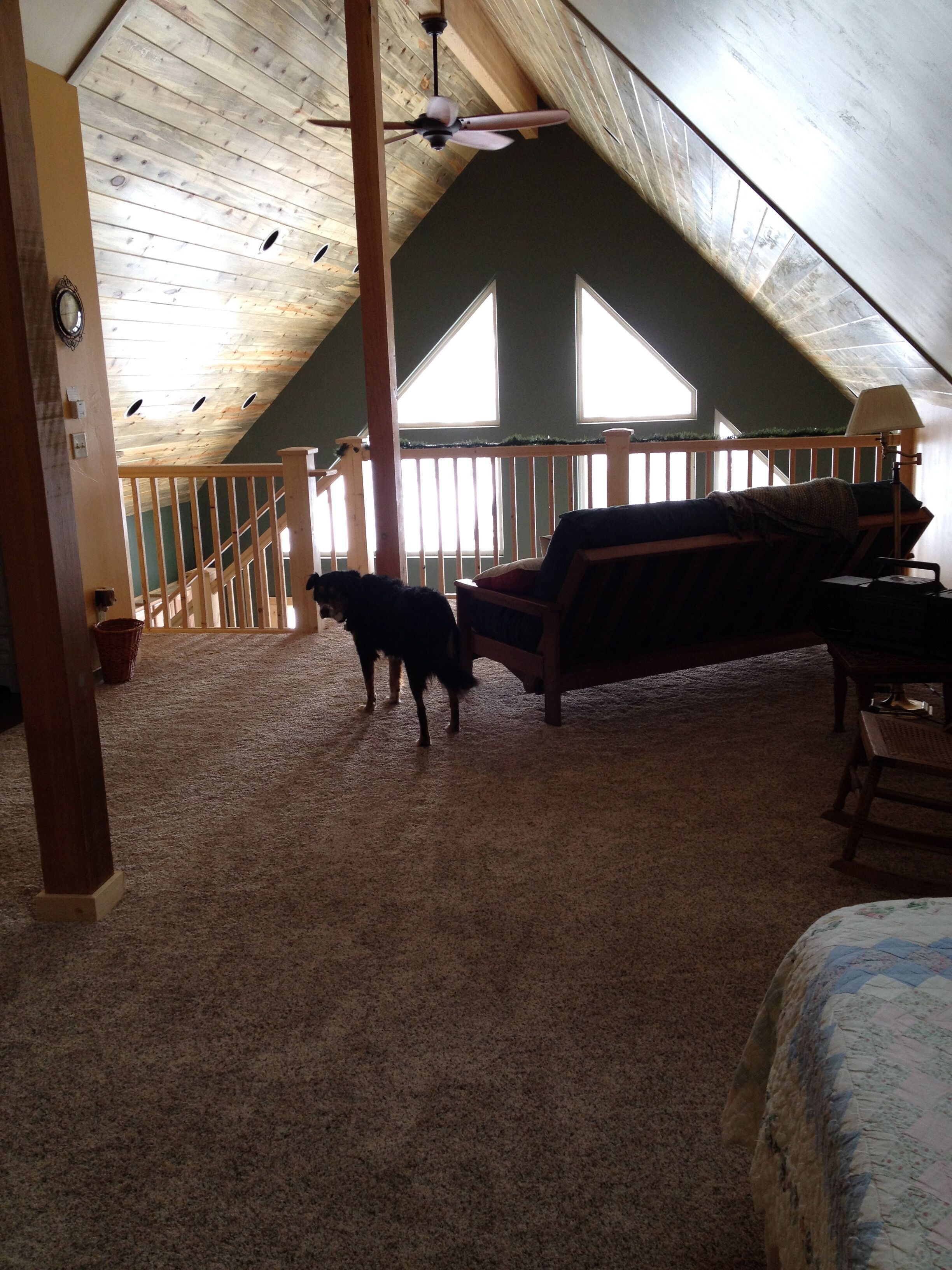An attic room with a sloped ceiling, wooden beams, a ceiling fan, a dog, a couch, and a window.