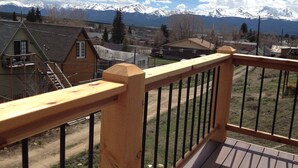 A wooden deck with a metal railing overlooking a residential area with houses and snow-capped mountains in the background.