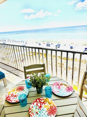 A table set with colorful plates and blue glassware on a balcony overlooking a beach with umbrellas. -  Seain is Believin / Beachfront Condo with Large Balcony/Conveniently Located (Gulf Shores)
