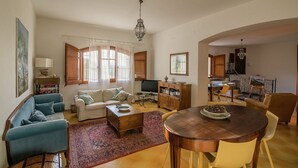 A spacious living room with a sofa, coffee table, and dining table. There is a television, a bookshelf, and a kitchen area in the background. - Charming Villa by the sea, wifi and cooking class free! (Termini Imerese)