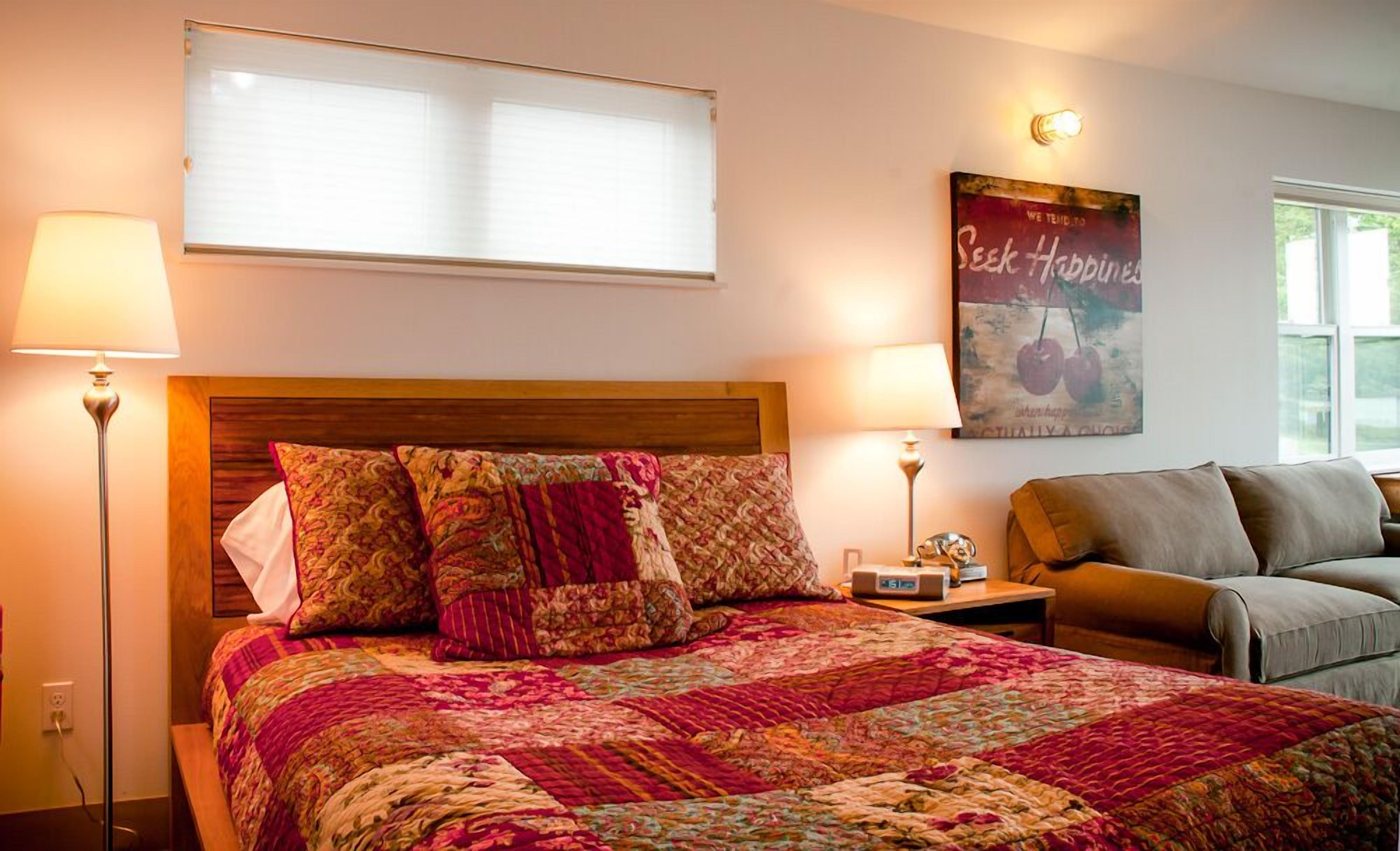 A bedroom with a large bed, two bedside lamps, a bedside table with a radio, a sofa, a window with blinds, and a poster on the wall.