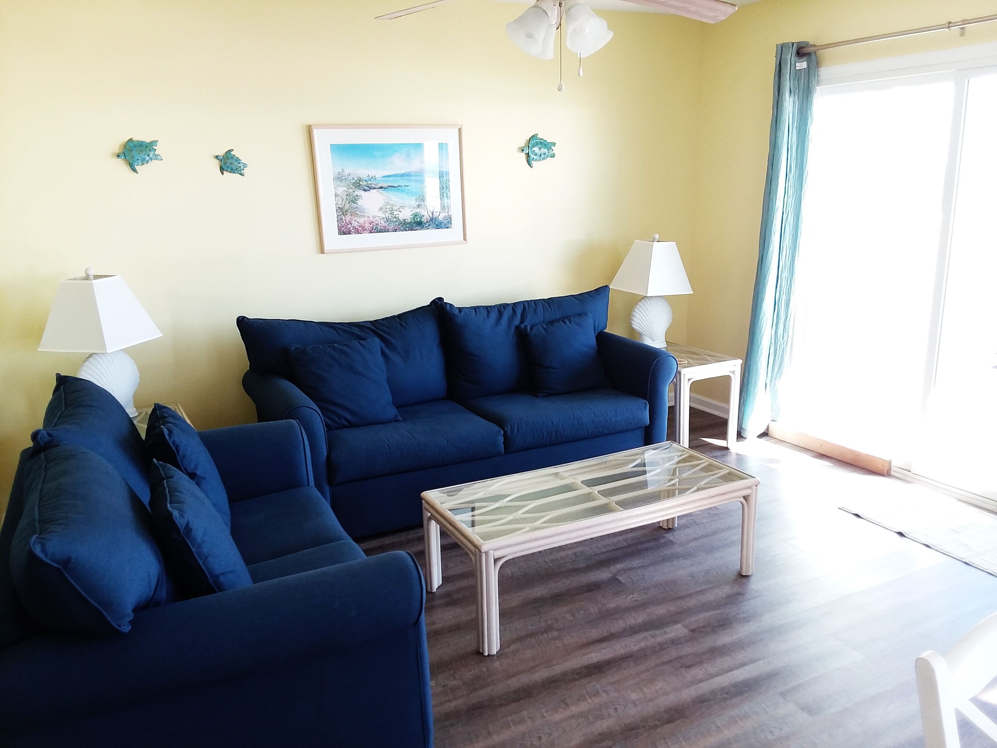 Un salon avec un canapé bleu, une table basse en verre et un tableau représentant une scène de plage. Il y a deux lampes sur des tables d’appoint et un ventilateur de plafond au-dessus.