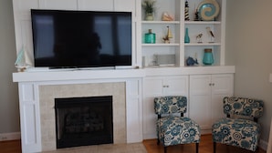 Uma sala de estar com lareira, uma televisão de tela plana montada acima, duas poltronas estampadas e um aparador branco com objetos decorativos.