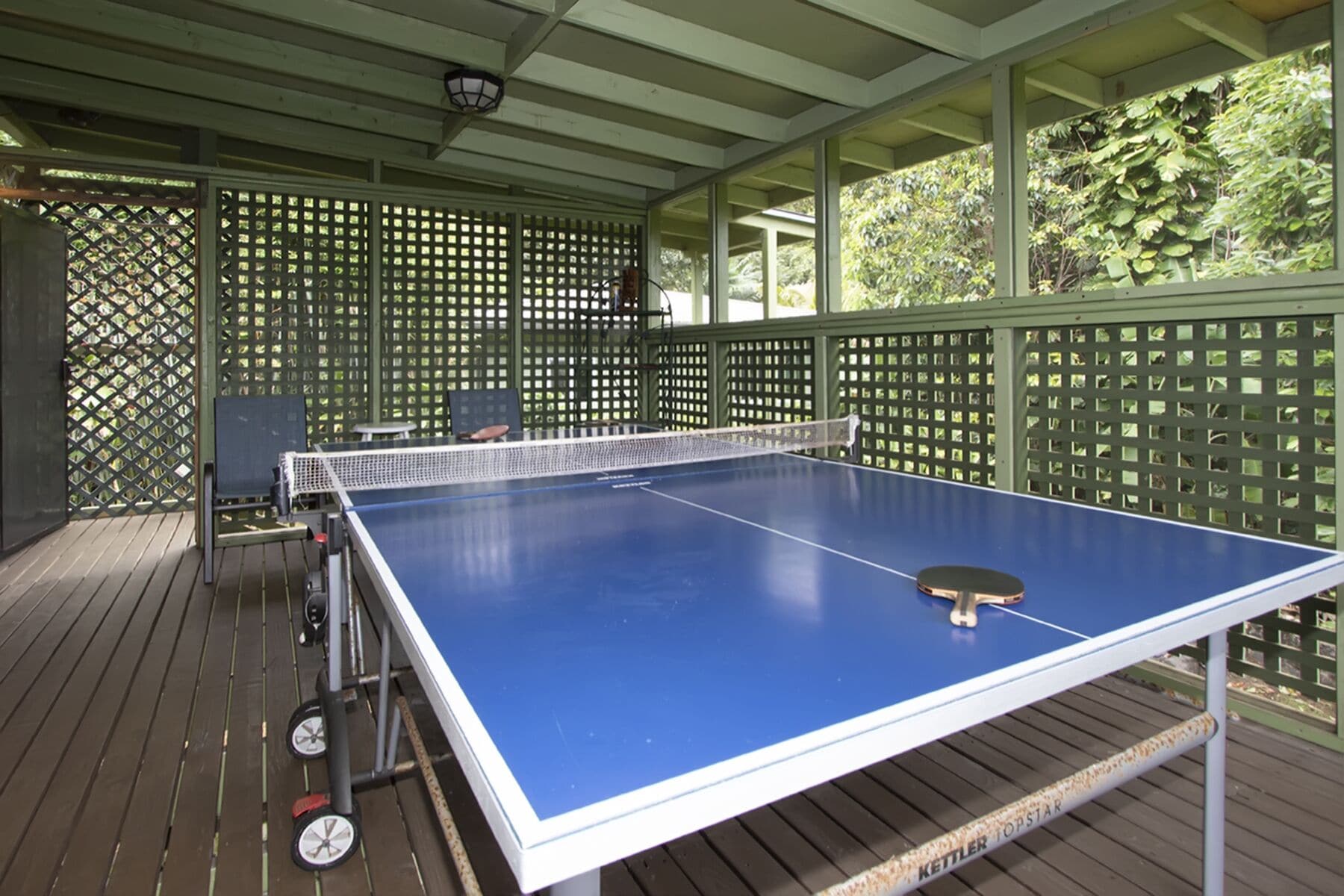 A ping pong table with a net in an outdoor covered area with wooden flooring and green lattice walls.