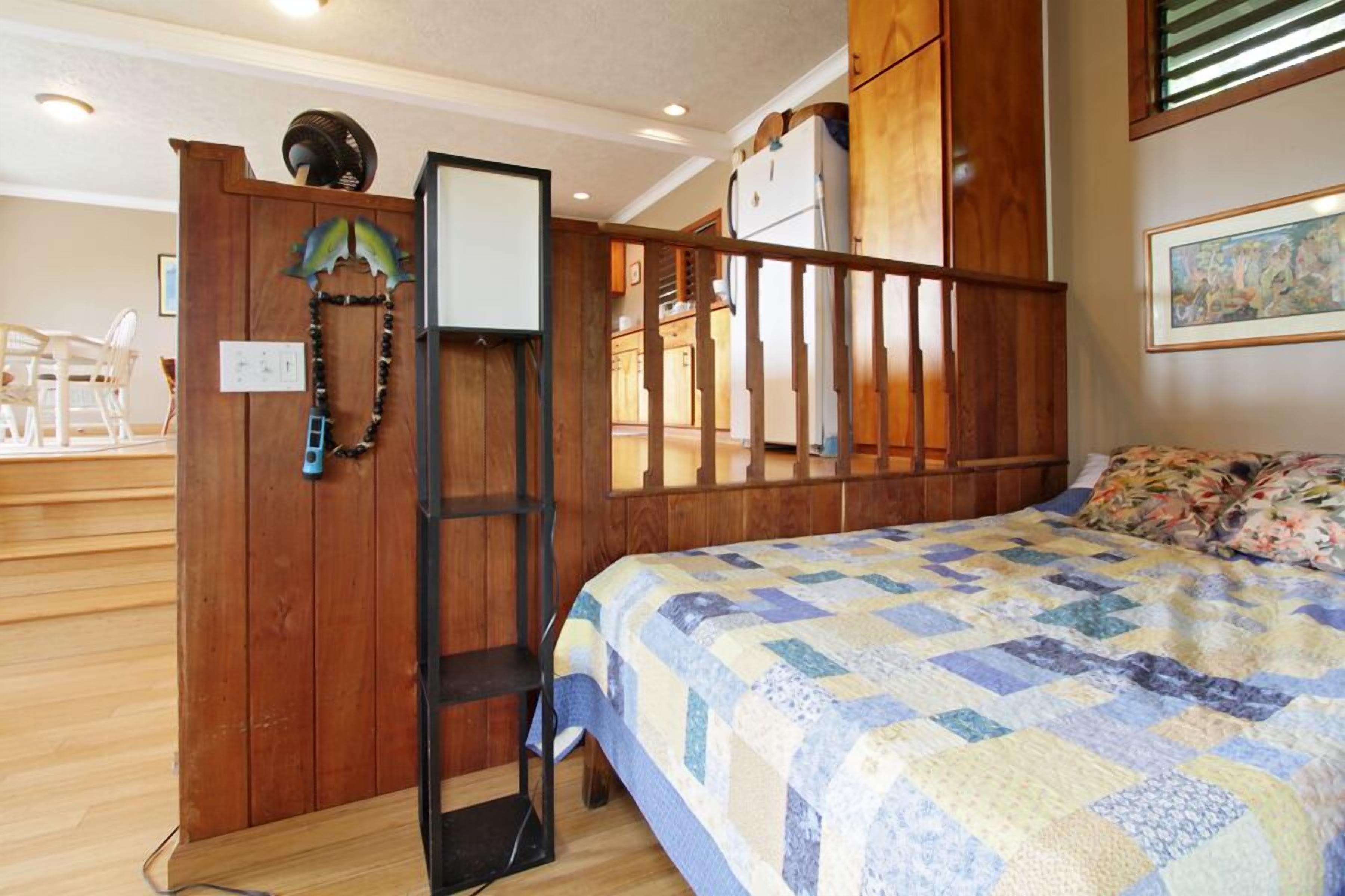 A bedroom with a wooden bed frame, a quilt, and a view of the kitchen through a wooden partition.