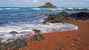 A beach with red sand, scattered rocks, and a distant rocky island. - Fisher's Hana Bay Hale, Across from Hana Bay (Kahului)