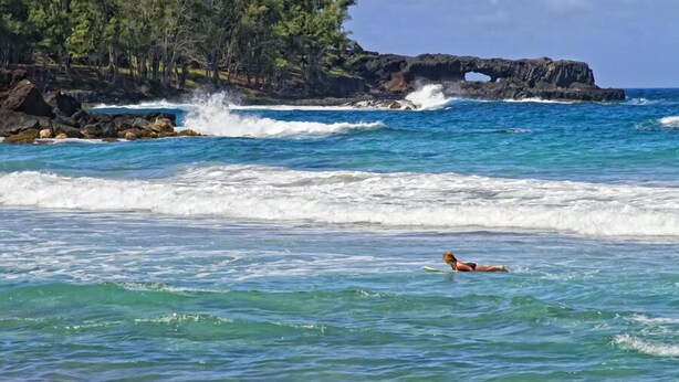 A beach with clear blue water, a person swimming, and a rocky cliff with a cave-like formation. - Fisher's Hana Bay Hale, Across from Hana Bay (Kahului)
