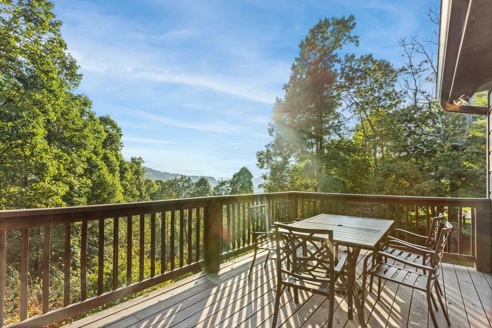 A wooden deck with a table and chairs, surrounded by a railing, overlooking a forested area with trees and a distant mountain range.