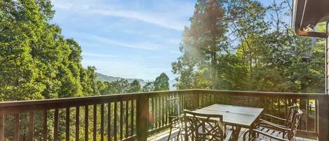 A wooden deck with a table and chairs, surrounded by a railing, overlooking a forested area with trees and a distant mountain range.