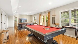 A pool table in a room with wooden floors, a kitchen area, and large windows with blinds. - Mountain Cabin On Quiet Property (Boone)