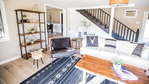 Een moderne woonkamer met een witte bank, een zwarte leren fauteuil, een houten salontafel en een tapijt met een patroon. Er is een trap met een zwarte leuning die naar een verdieping leidt.