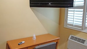 Une pièce avec un téléviseur fixé au mur, un bureau en bois avec une chaise, une télécommande et une fenêtre avec des stores.